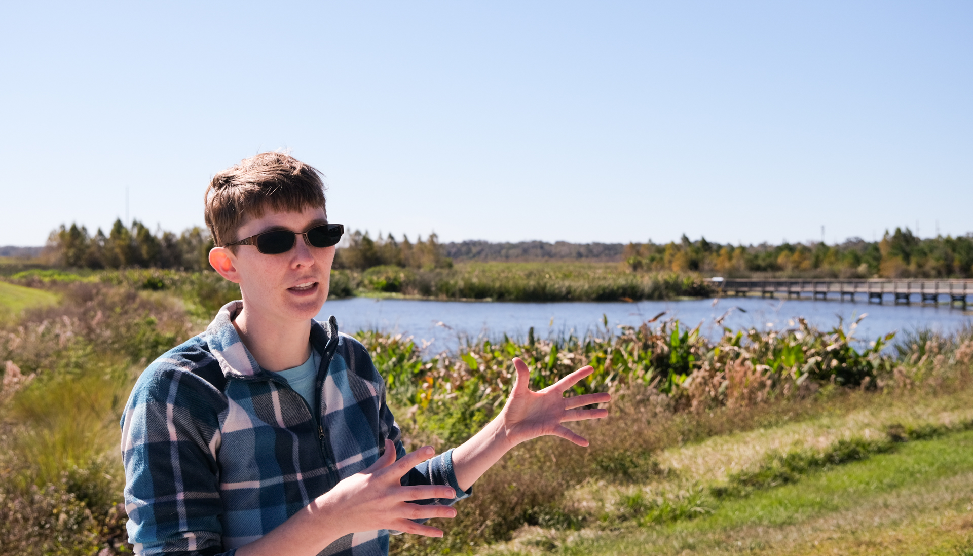 A person with short hair, glasses, and a plaid flannel jacket gestures with their hands while speaking. There is a body of water behind them, with a boardwalk and water plants visible.