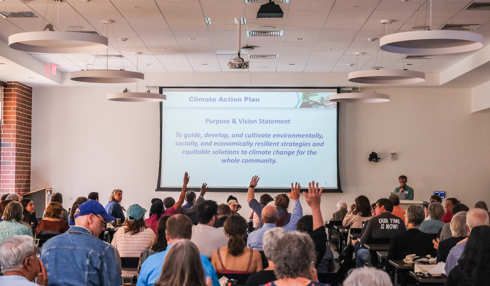 A man at a podium stands next to a projector screen with the words, "Climate Action Plan: Purpose & Vision Statement. To guide, develop, and cultivate environmentally, socially, and economically resilient strategies and equitable solutions to climate change for the whole community." There is a crowd of several dozen people in front of him. Some of their hands are raised.