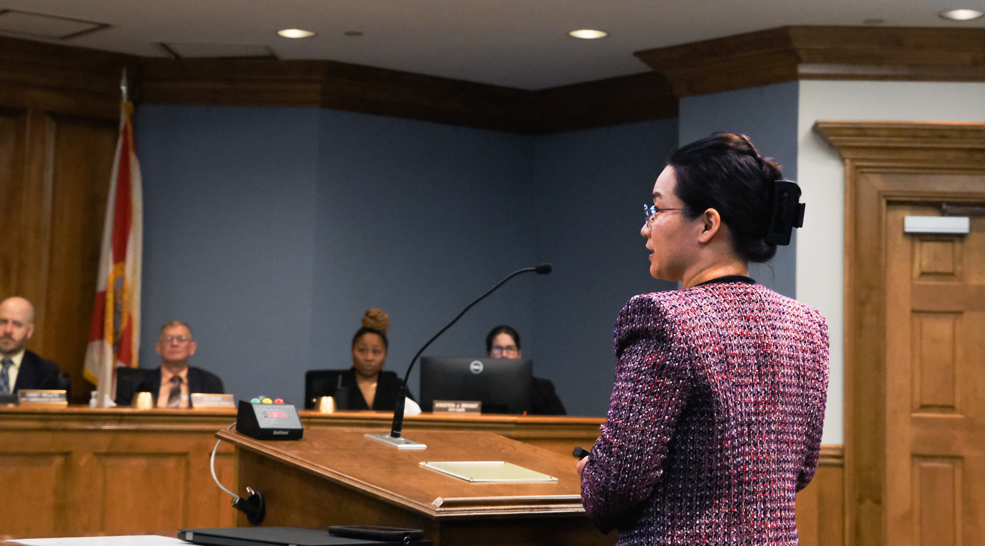 a woman wearing glasses, a black hair clup, and red tweed jacket stands at a podium in front of an assorted group of people in suits and formal work wear behind a city council bench, each has a name tag in front of them. There is a draped Florida state flag behind the people at the city council bench.