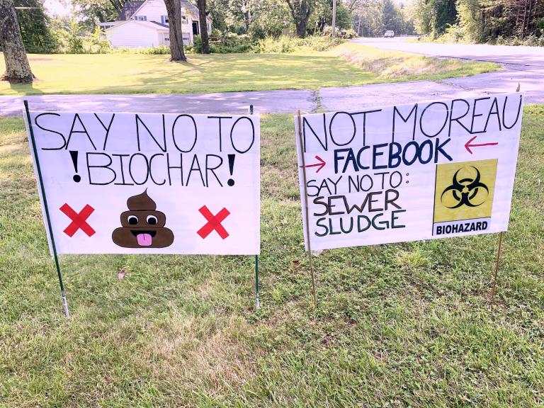signs that say 'say no to biochar' and 'not moreau' on a lawn. The biochar sign has a poop emoji drawn on it