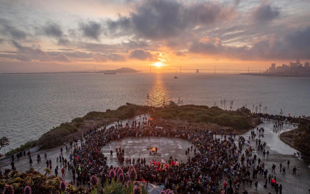 An aerial photo shows a crowd of people forming a large circle around a bonfire at the tip of an island, as the sun rises in the background