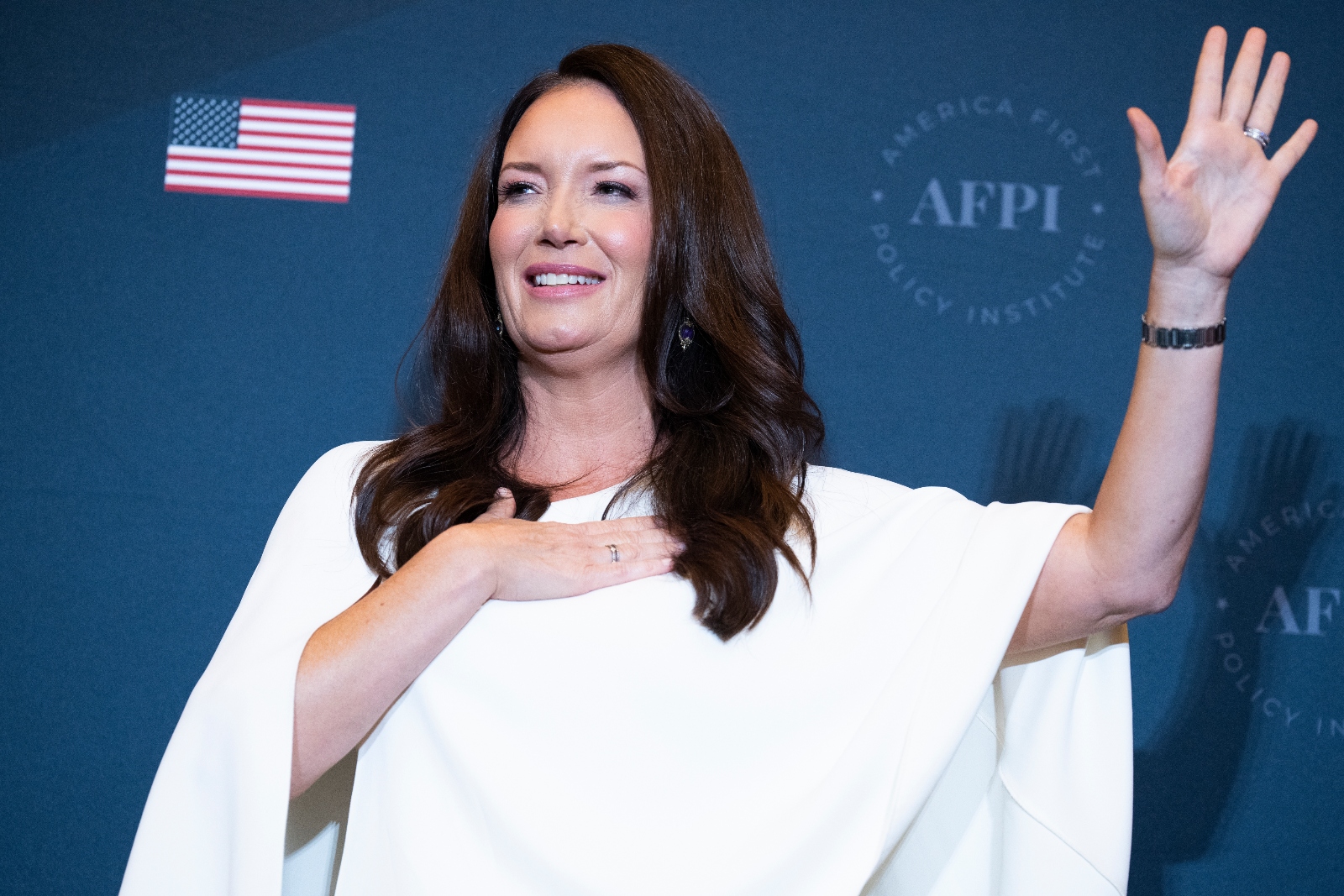 A white woman with long dark hair, wearing a flowing white blouse, stands with her hand over her heart at a podium