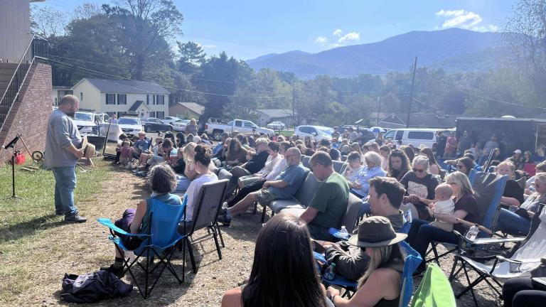 Outside a brick building, under a blue sky with mountains in the background, a man in a grey shirt and jeans holding a hat stands in front of a group of people seated in folding chairs.