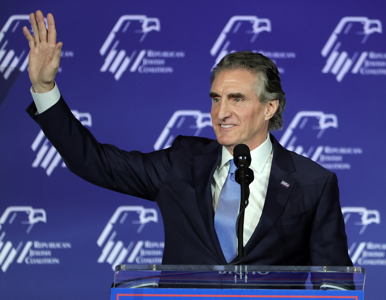 A man in a suit with graying hair waves from a podium