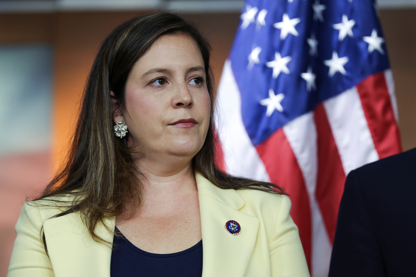 Closeup a white woman with long dark hair wearing a black shirt and yellow blazer standing with the American flag in the background