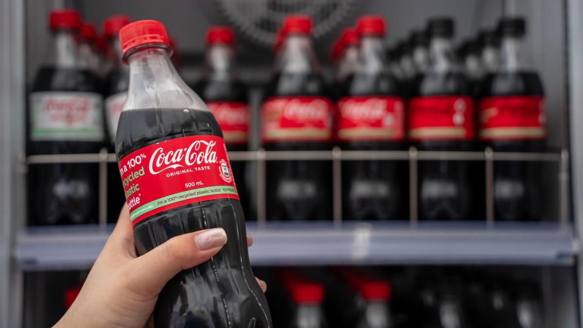 Close-up of a hand holding a Coca-Cola bottle, with a shelf of more bottles in the background.