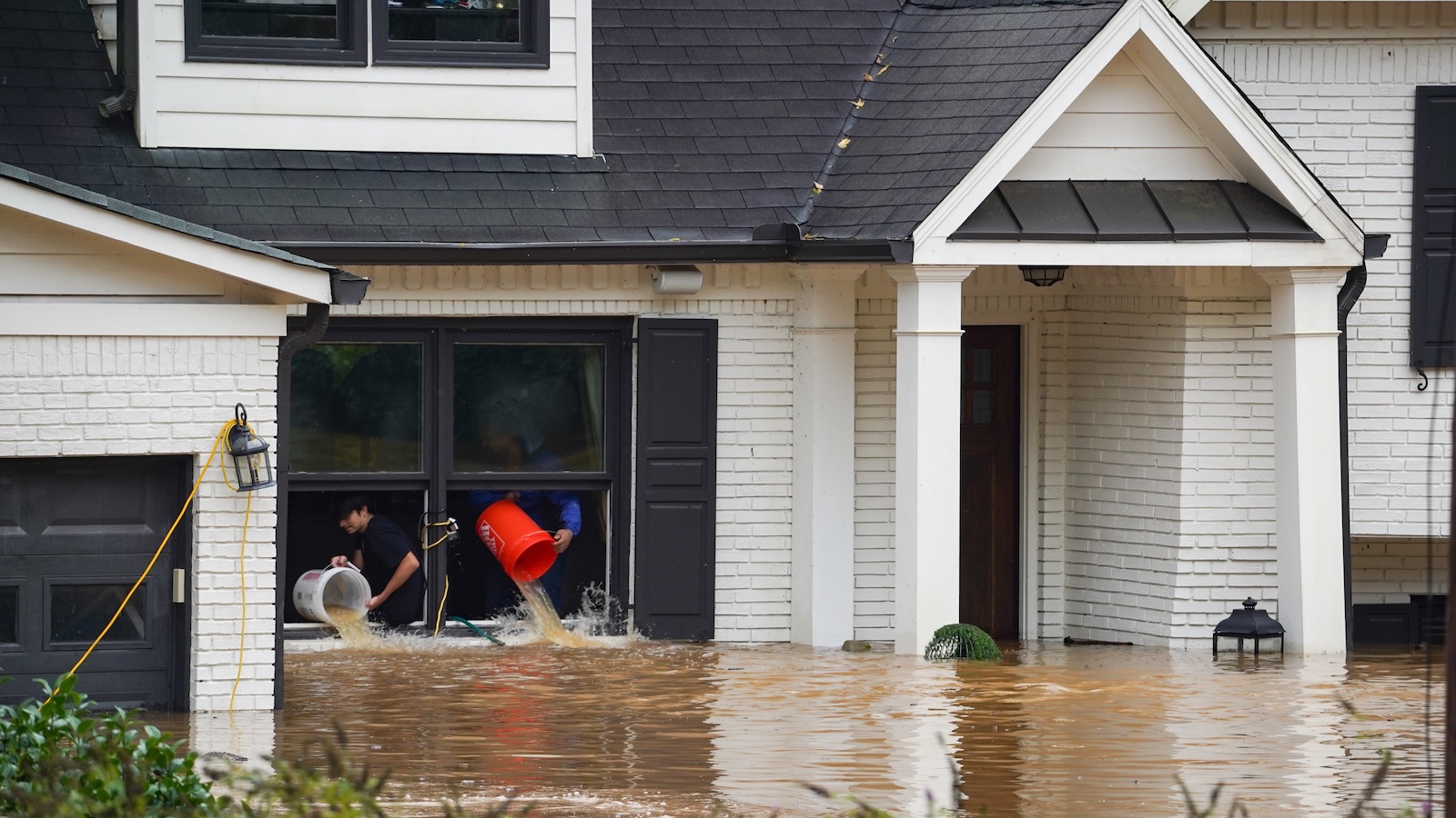 Two homeowners use buckets to bail water through the window of their flooded home.