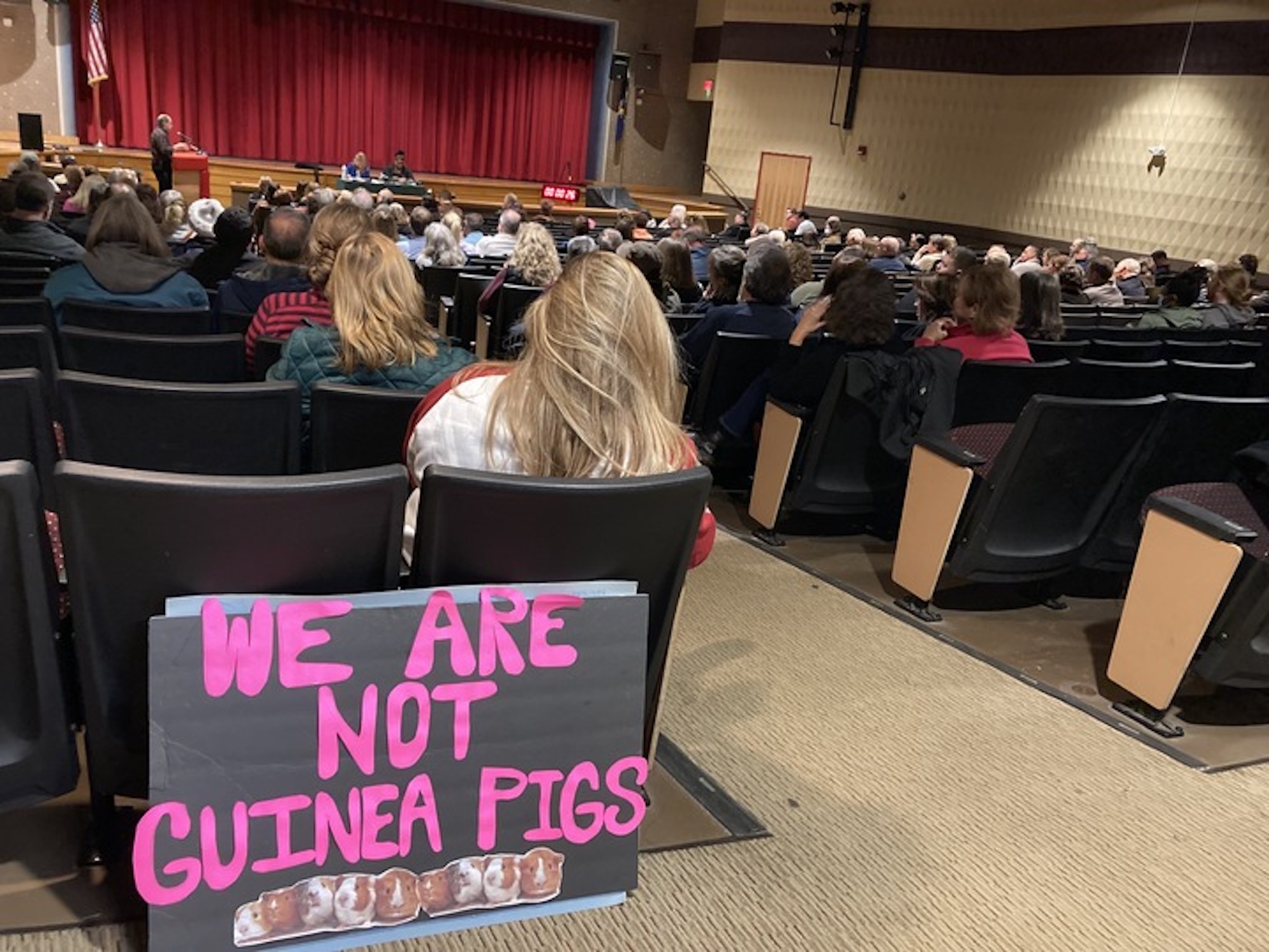 A group of people sit in an auditorium with a sign that says 'we are not guinea pigs'