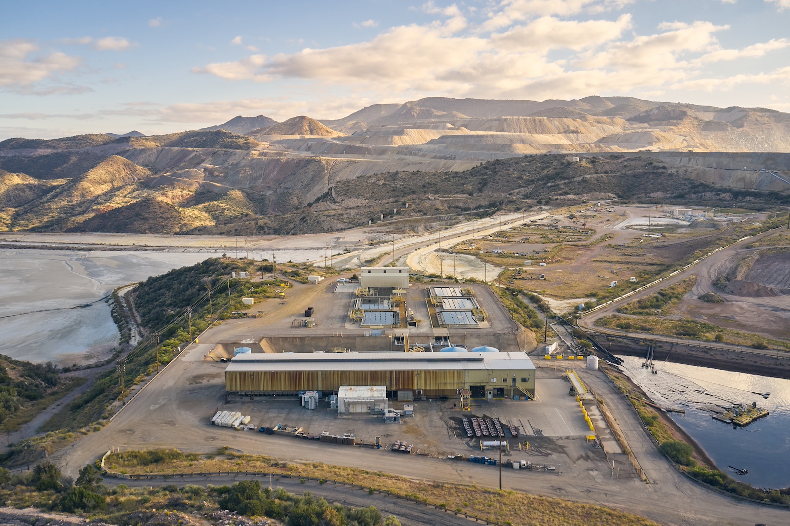 An aerial shot of an industrial facility, with a pond to the right and tan mountains and a blue sky with wispy clouds in the background