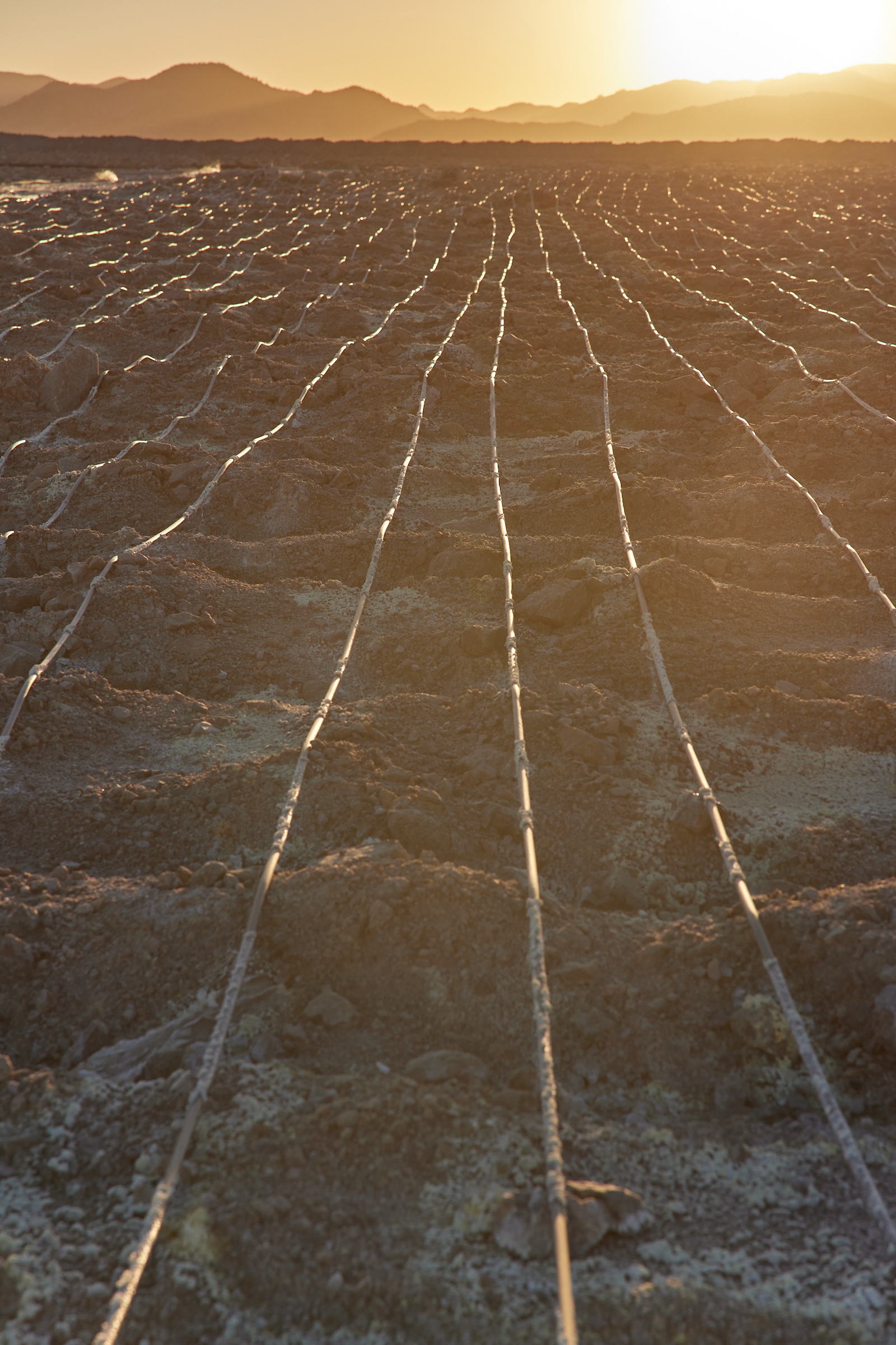 Long tubes run toward the horizon across piles of crushed rock, as the sun sets on mountains in the background