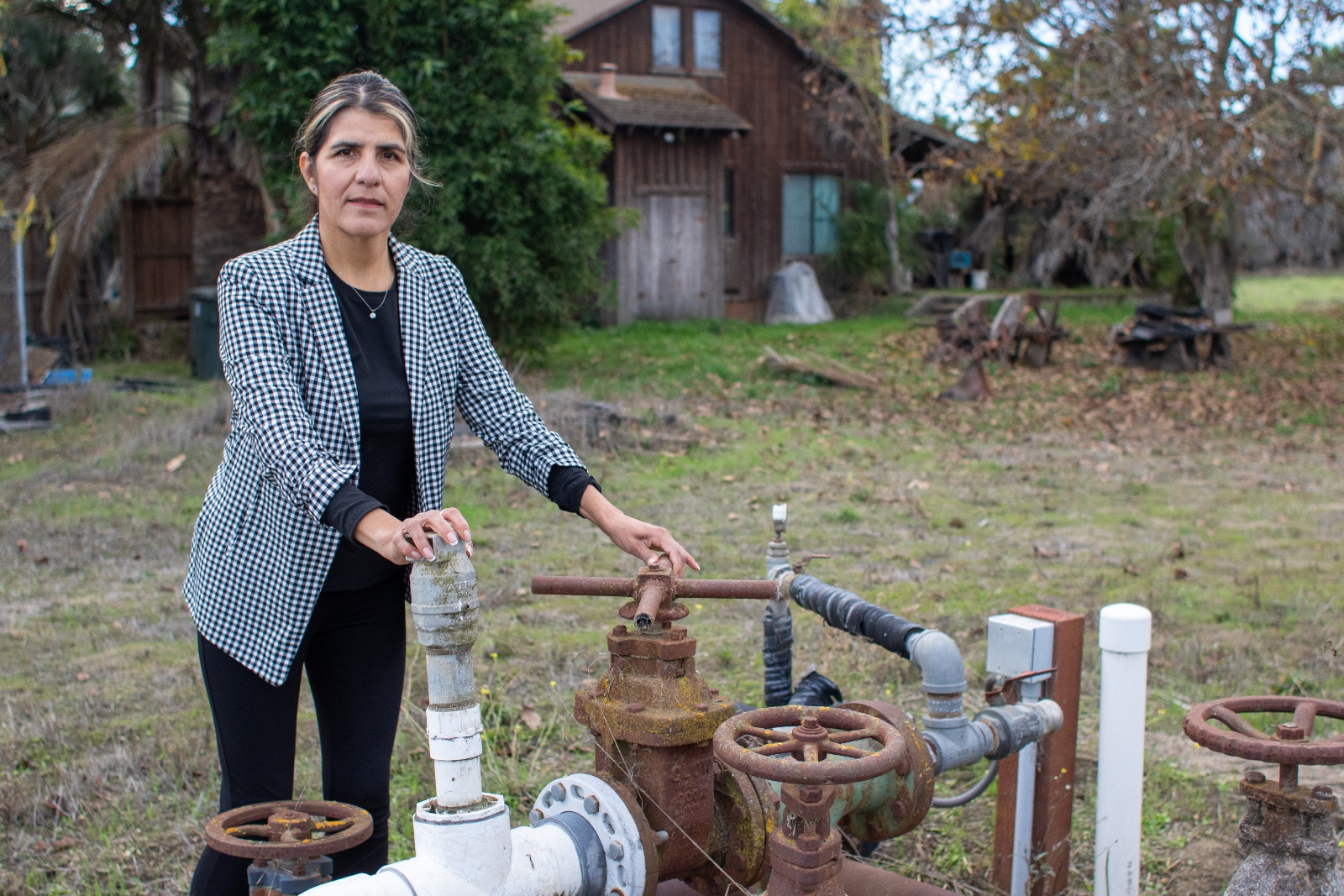 A woman wearing a blazer stands next to a water well with a farmhouse in the background