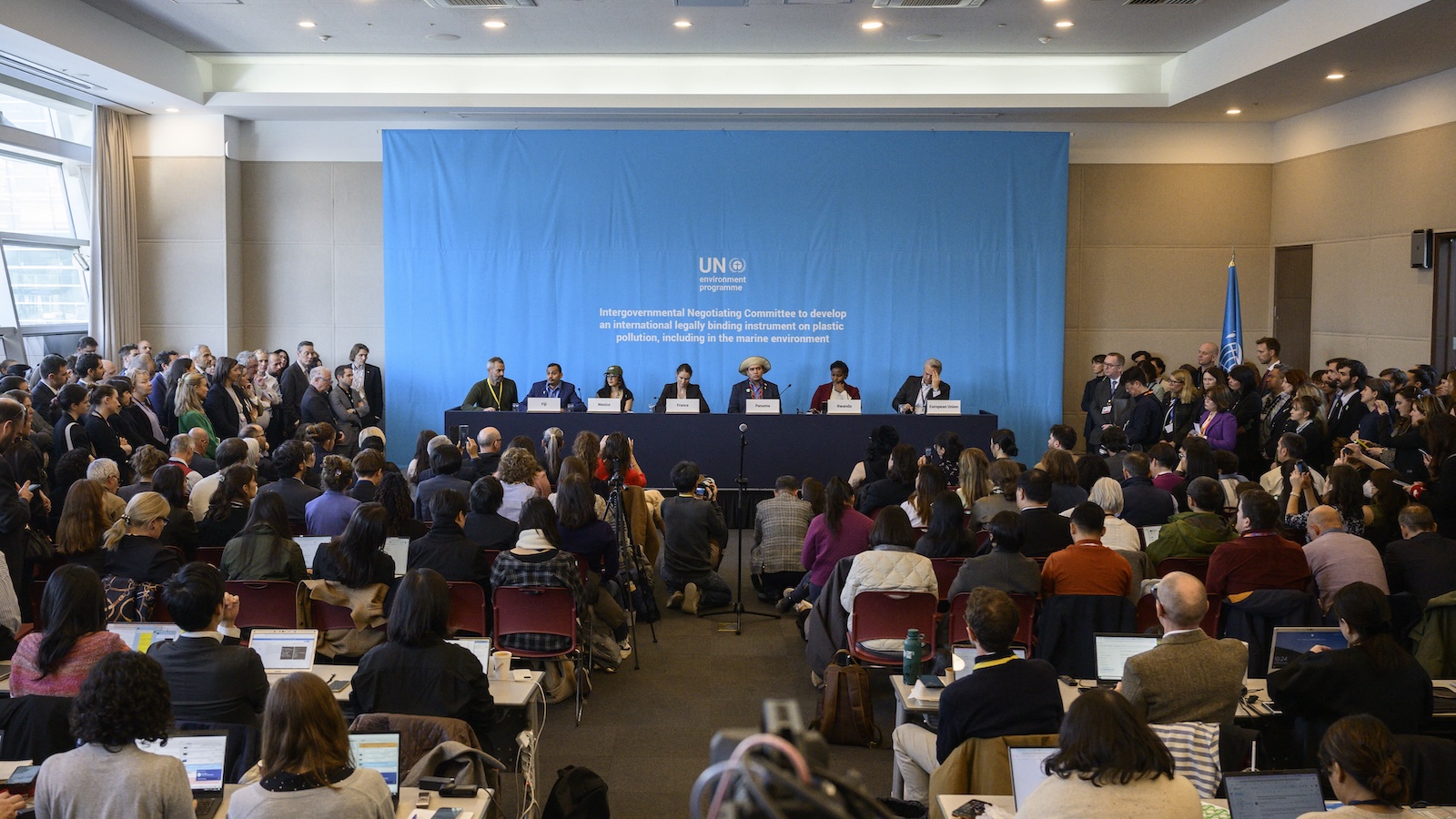 A crowded room of seated people facing a row of diplomats seated at a table, with a blue backdrop.