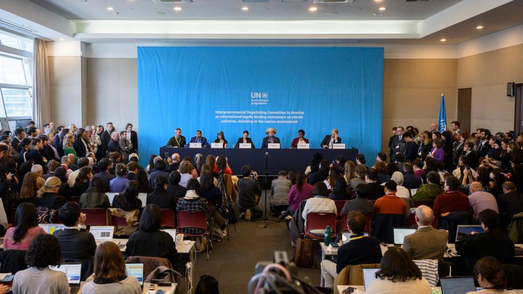 A crowded room of seated people facing a row of diplomats seated at a table, with a blue backdrop.