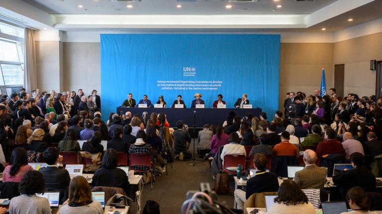 A crowded room of seated people facing a row of diplomats seated at a table, with a blue backdrop.