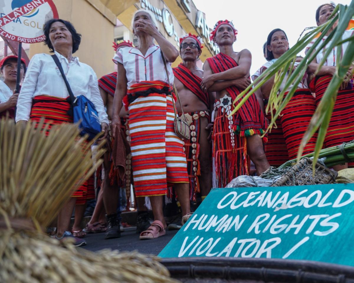 a group of people in red and white clothing standing near a sign that says 'oceanagold human rights violator!'