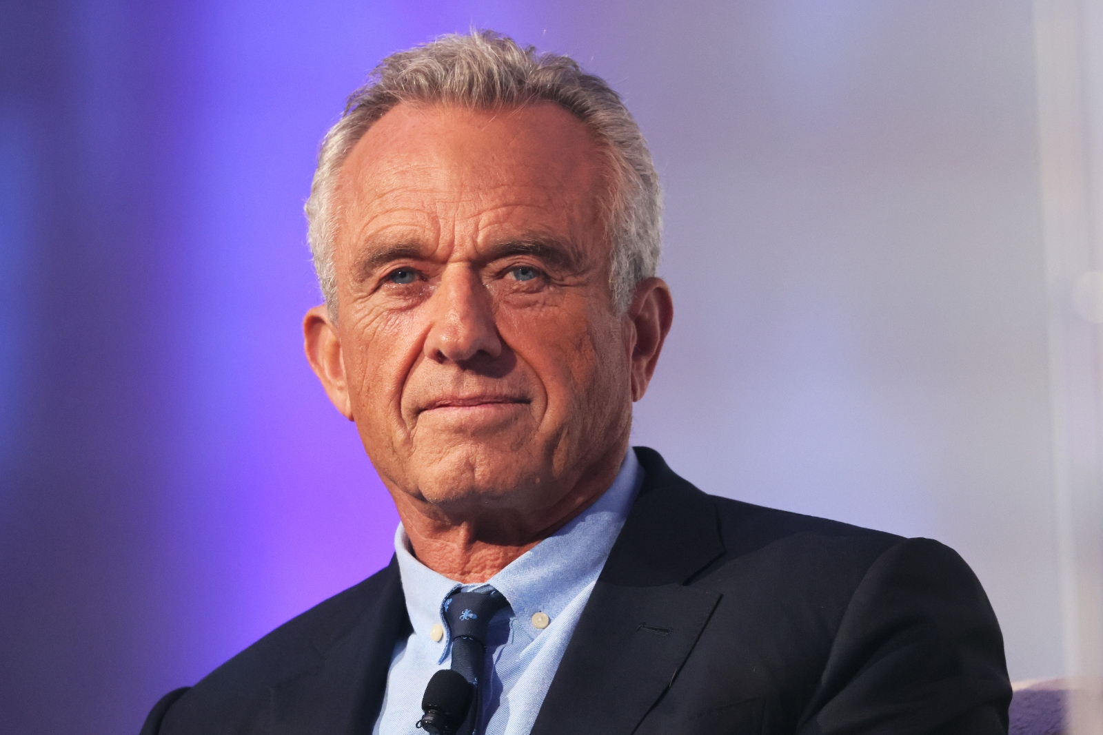 A closeup photo of a white man with gray hair in his 70s, wearing a suit and tie in front of a purple background