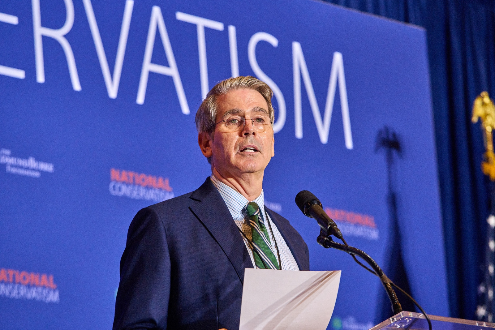 A middle-age white man wearing glasses and a suit and tie speaks from a podium at a conference