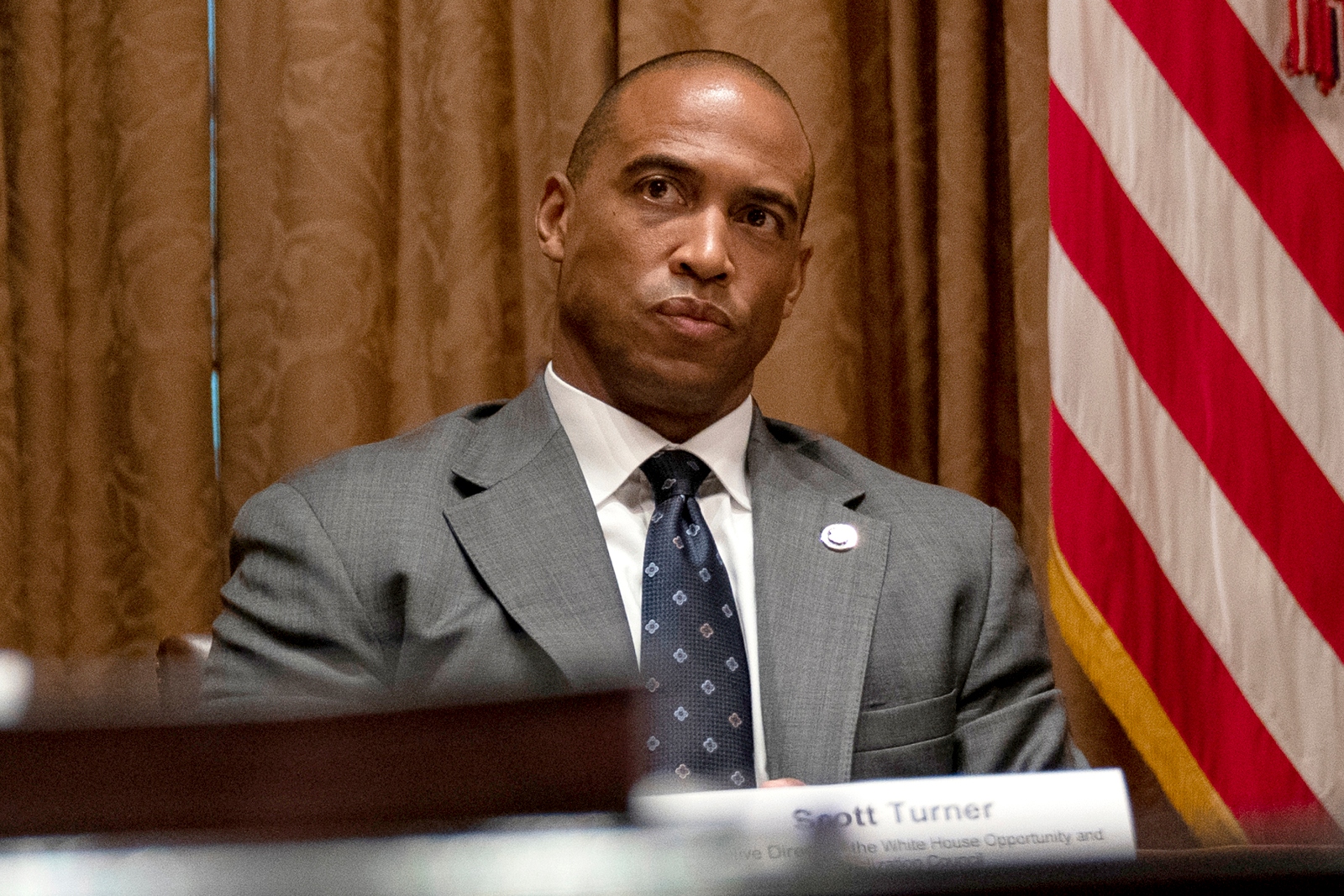 A bald Black man in a gray suit and tie sits on a dais next to an American flag