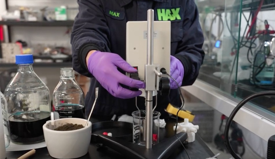 The torso of a person wearing a navy blue sweatshirt with the neon green text 'Max Hax' on it. The person is wearing purple disposable gloves and adjusting a metal piece of equipment next to some bottles full of dark blue liquid and a bowl full of dark powder