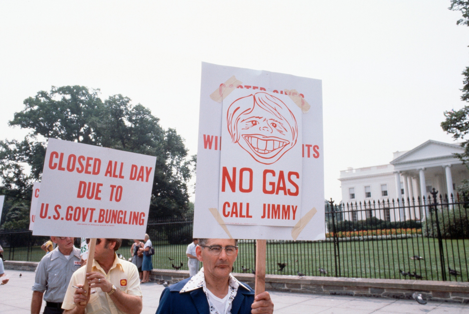 A color photograph of protesters marching outside the White House. One man carries a sign reading Closed All Day due to U.S. Government Bungling. Another carries a sign with a caricature of Jimmy Carter's face and the words No Gas Call Jimmy.