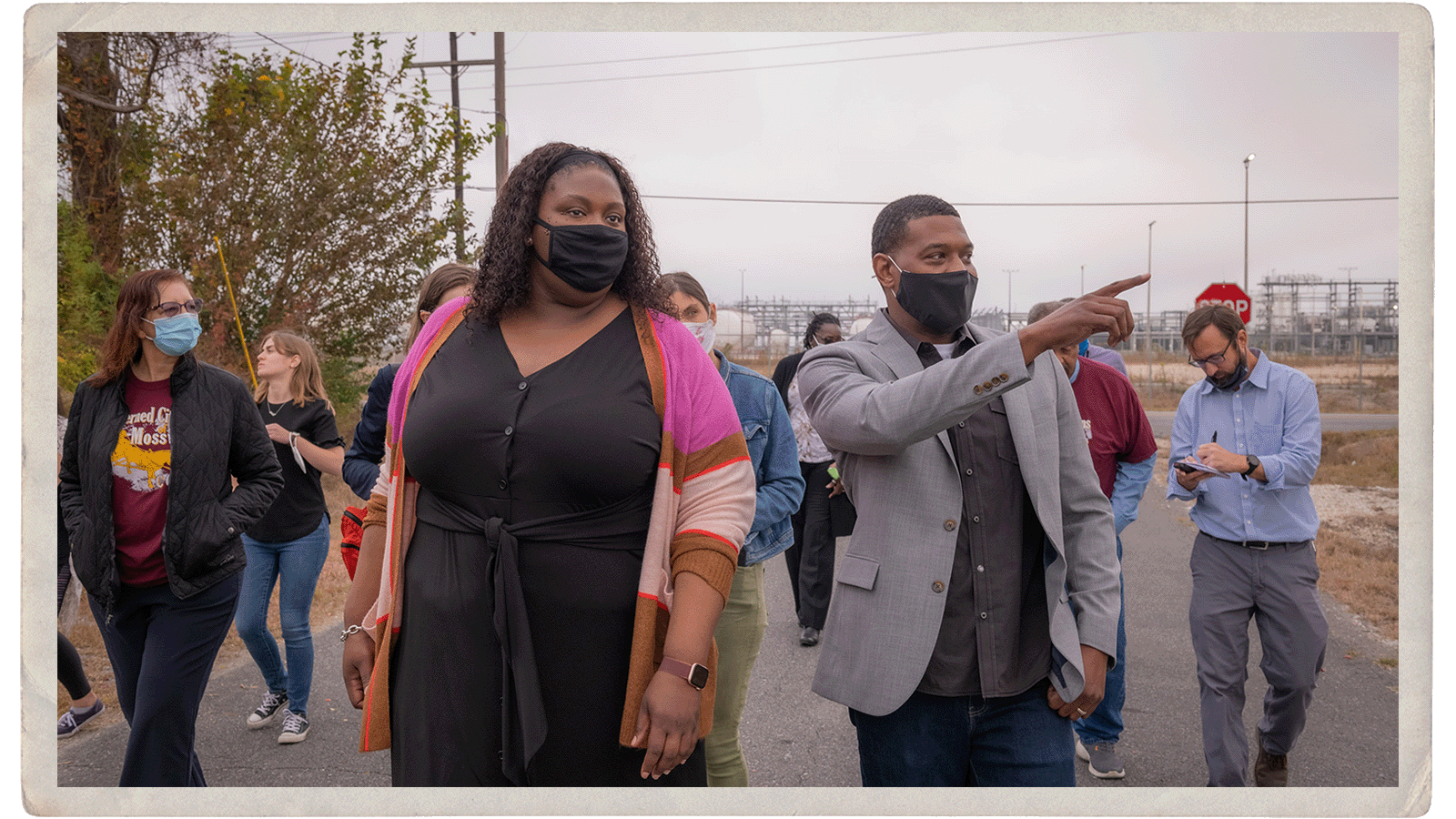 A group of people walk along a tree-lined street. One, Michael Regan, is pointing at something off-scene.