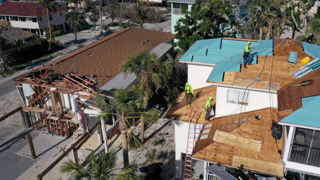 A group of workers in bright yellow stand on wood planks on a roof next to a building damaged by a hurricane