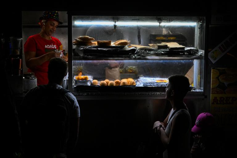 People on a dark street wait in line behind a food stand illuminated by a fluorescent light. The server wears a festive hat