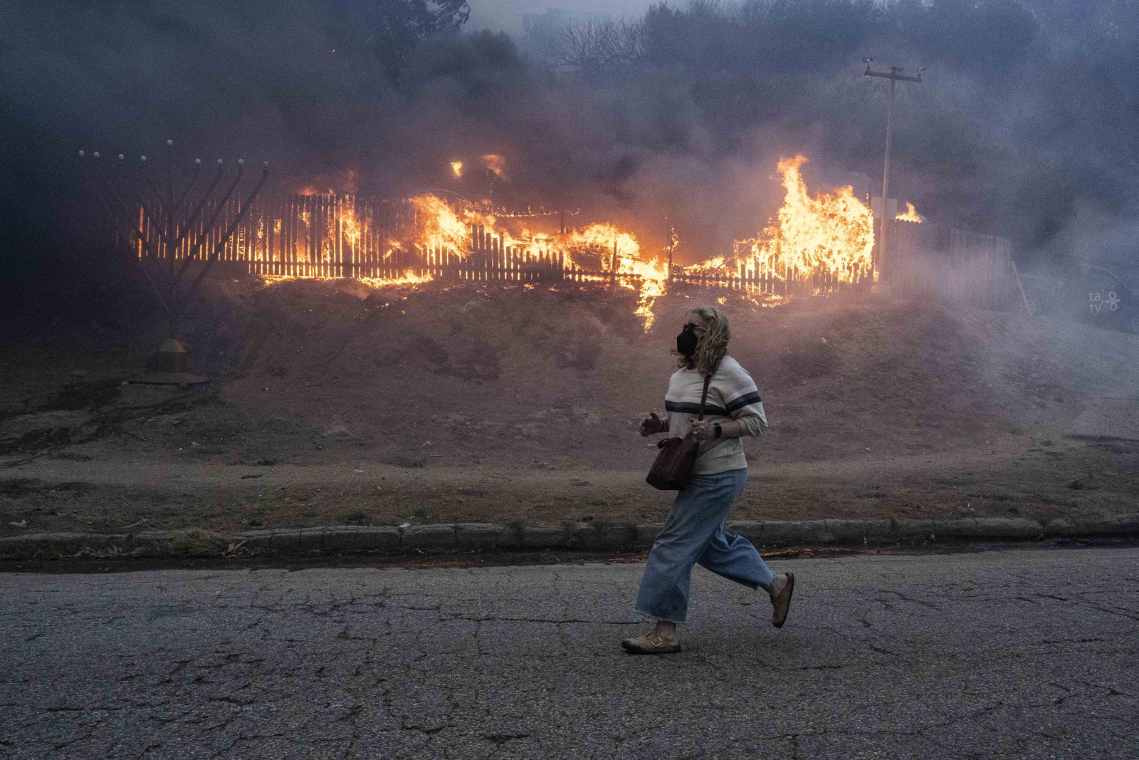 a woman with shoulder length blonde curly hair and baggy jeans runs by a flaming fence. she is wearing a black mask. her birkenstock clogs are streaked with soot.