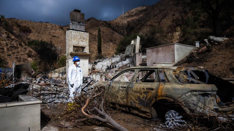 A man in a white jump suit and blue hard helmet stands in the remains of house that burned down and next to a burned out car