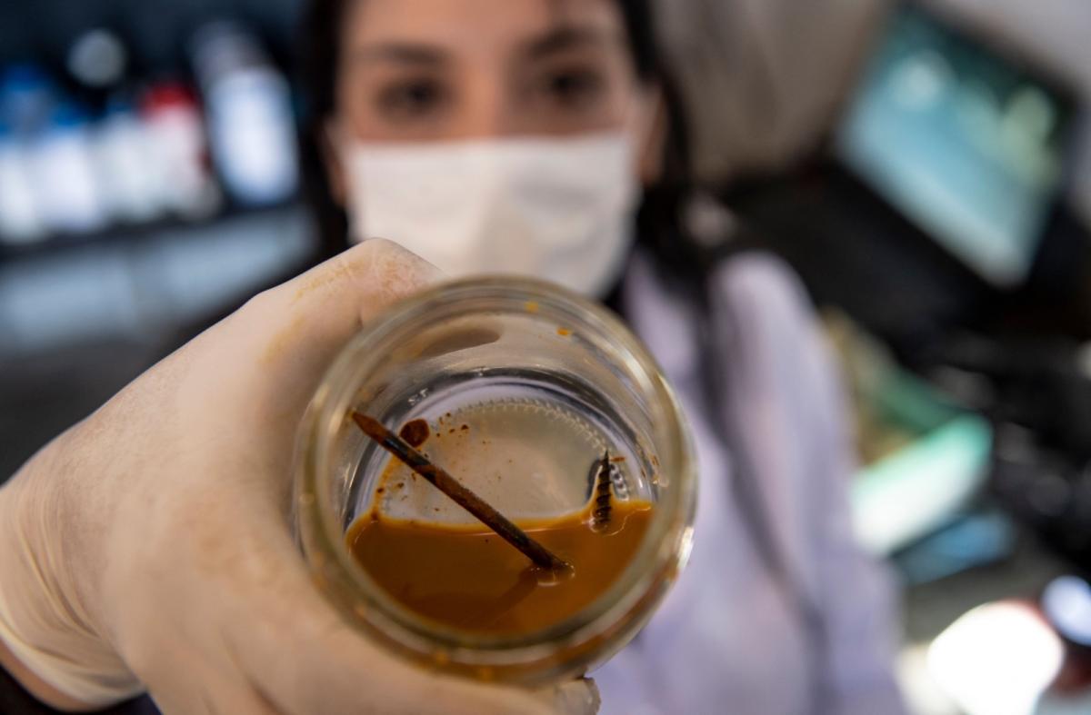 A photo of a woman in a mask and gloves holding up a glass jar with a nail submerged in a rust-colored liquid