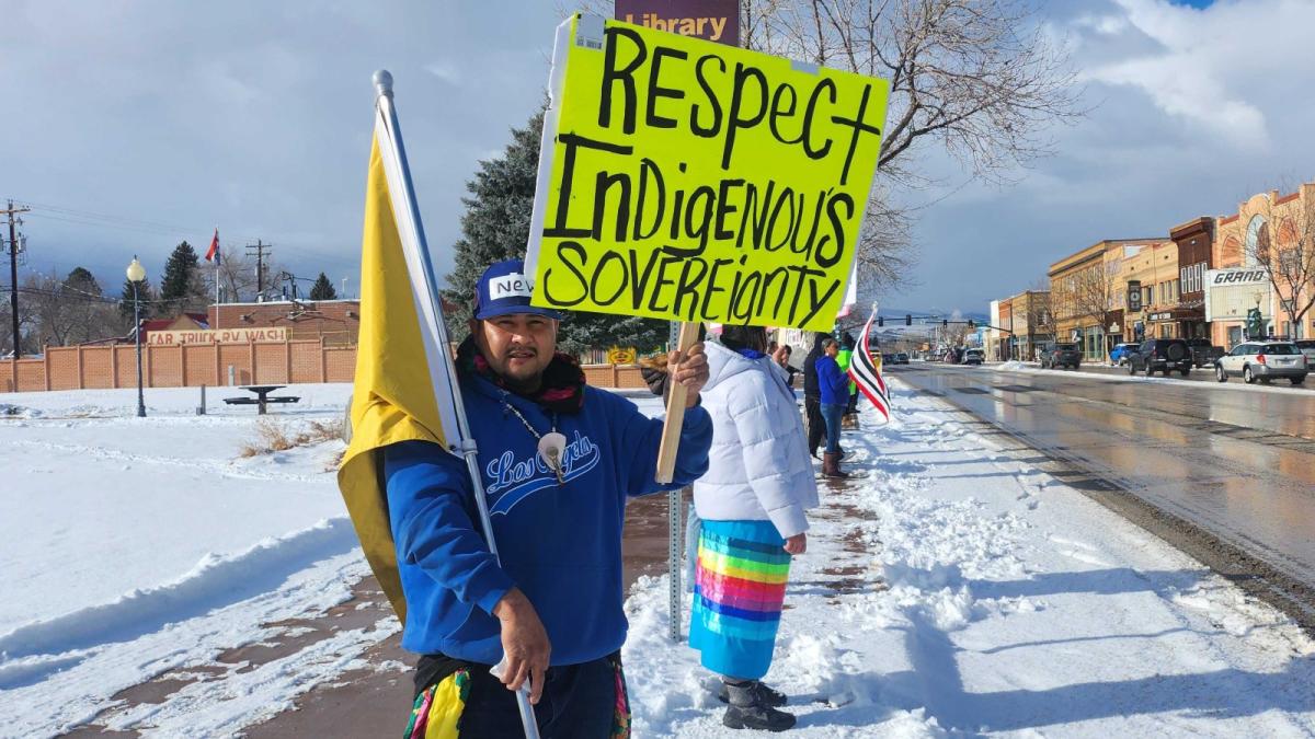 Man holds sign that says "Respect Indigenous Sovereignty" in one hand, and a Eastern Shoshone flag in the other on a winter day in protest.