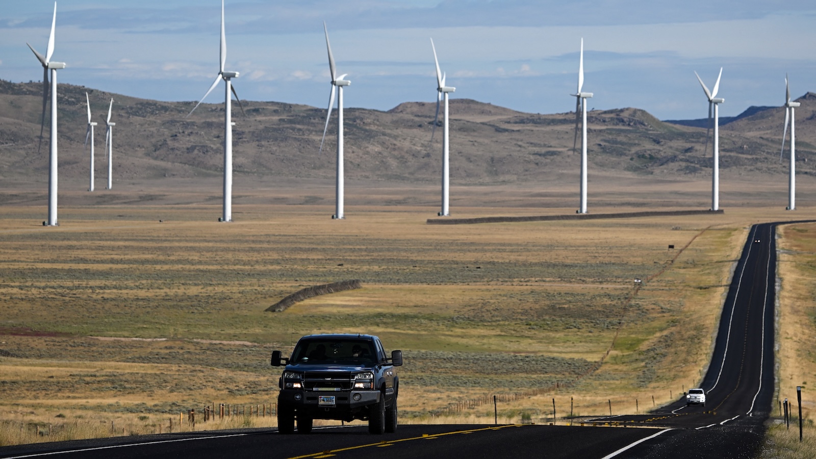 A pickup truck drives down a long straight highway with a row of wind turbines in the distance.