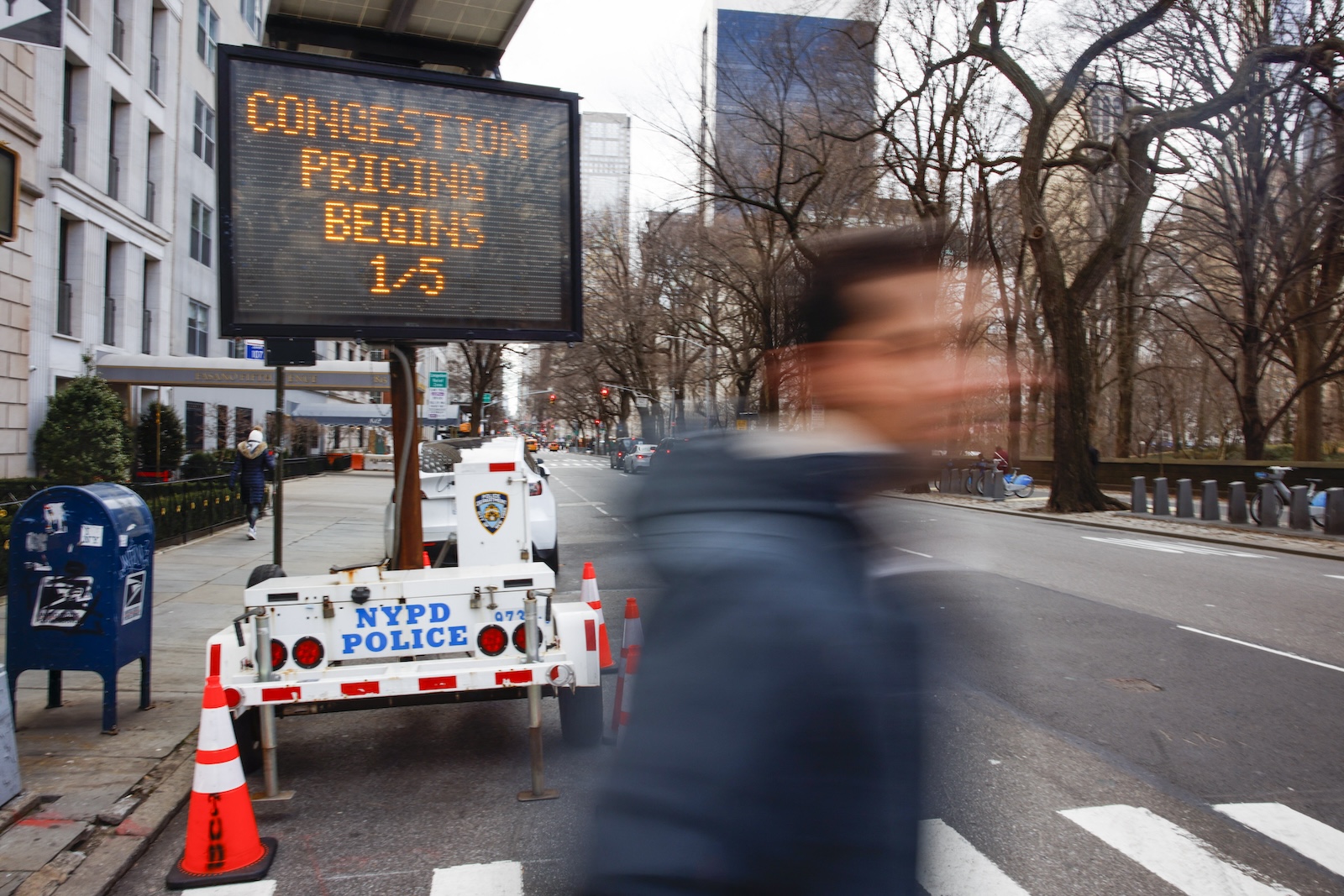 A man walks in front of a sign announcing the start of congestion pricing in New York City, his face blurred.