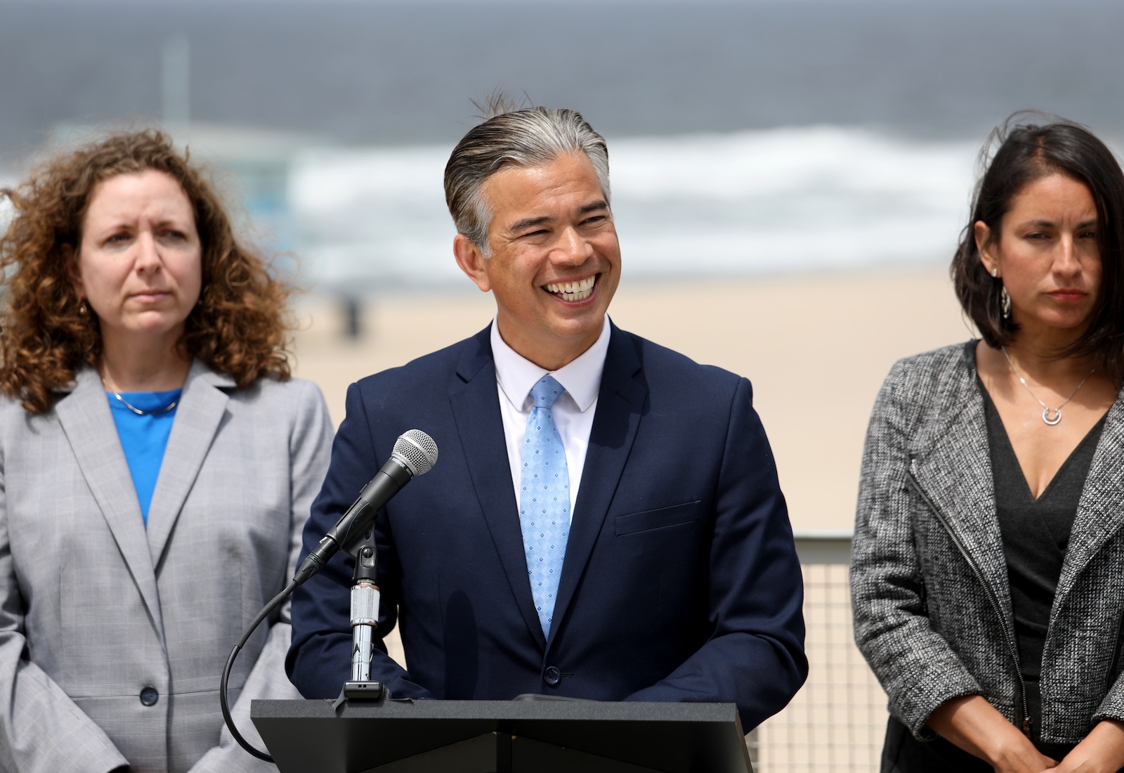 Rob Bonta stands at a podium with beach in the background, smiling at camera