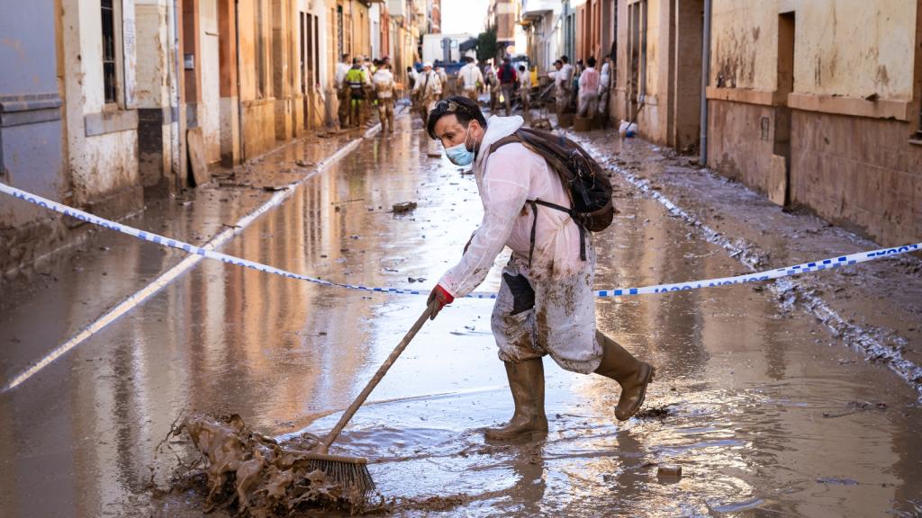 A man in a white jump suit and medical mask and boots sweeps a muddy, water filled street