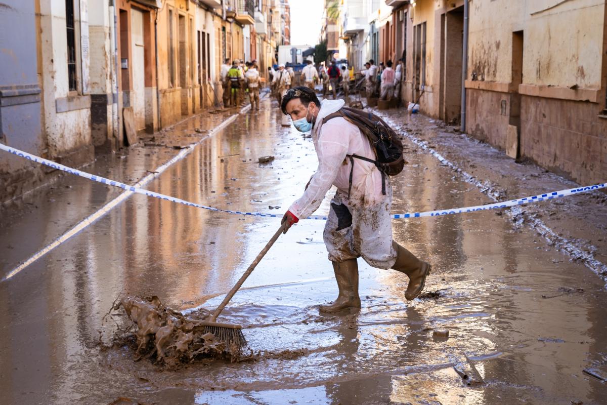 A man in a white jumpsuit and medical masks cleans a water logged street with a broom