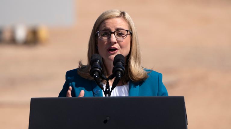 A blonde woman with glasses stands at a podium