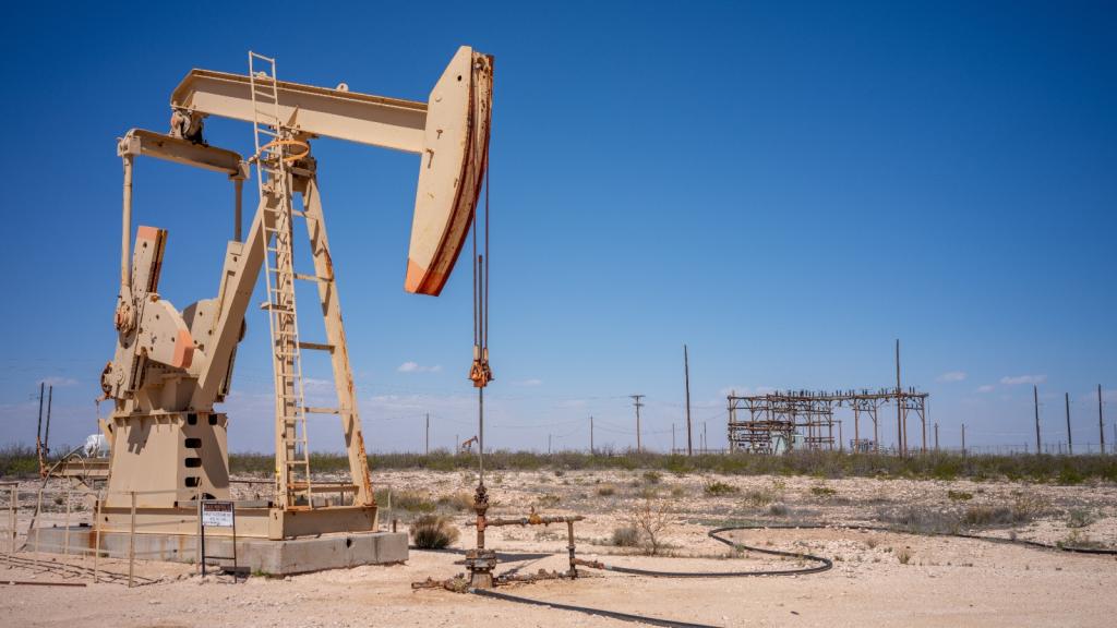 A giant beige pumpjack sits in the middle of the desert next to an industrial building