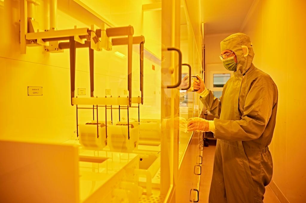 A person wearing a face mask, a hazmat suit, and gloves stands in front of a glass case filled with equipment. The entire scene is bathed in yellow light.