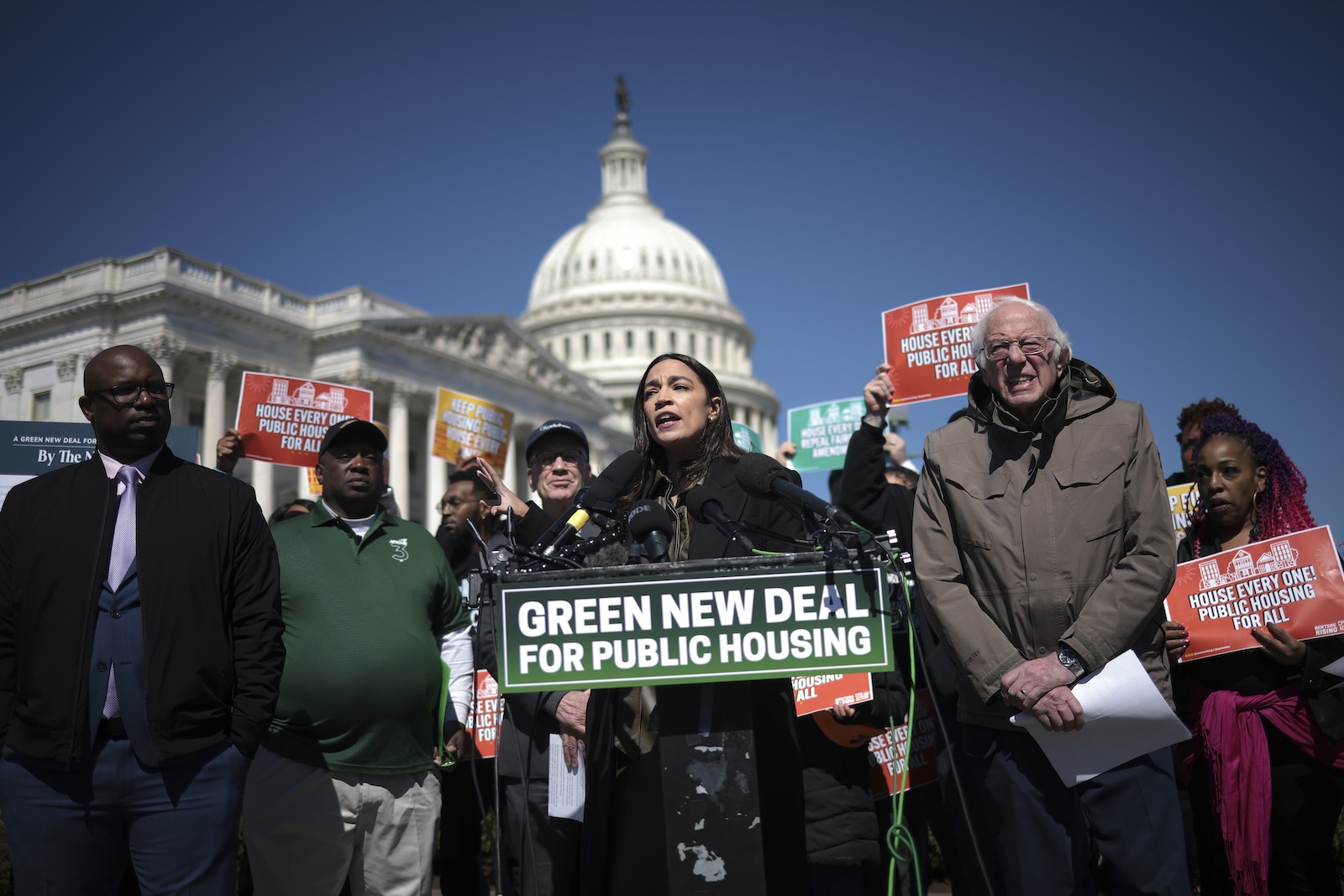 Congresswoman Alexandria Ocasio-Cortez delivers a speech behind a podium with the sign "Green New Deal for Public Housing", with Senator Bernie Sanders to her left and the White House in the background