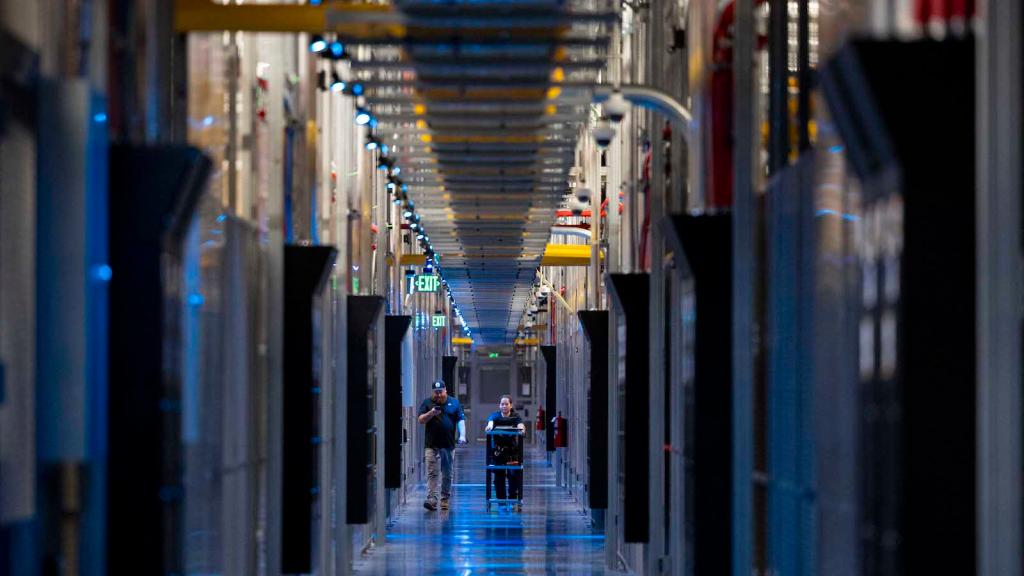 Two people walking down a hallway lit with blue lights and large looming computer towers.