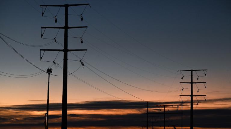 Big black electric structures with wires draped between them against a dark blue sky, with a pink sunset and dark clouds at the bottom