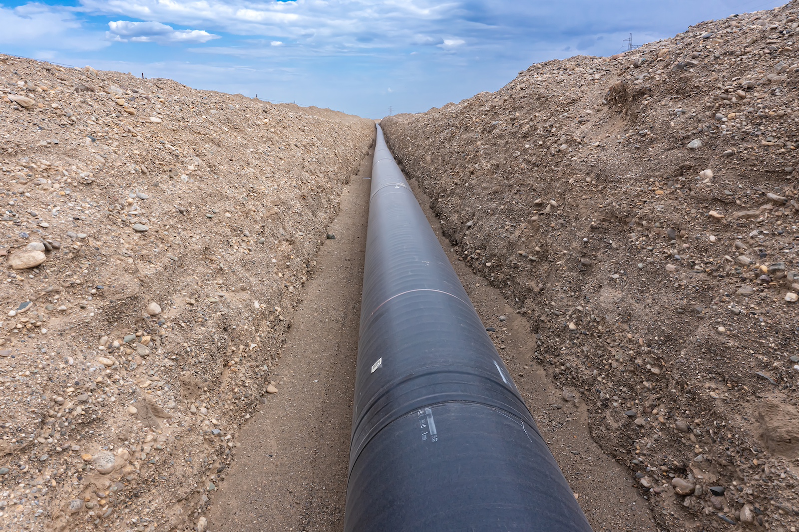 A black pipe runs toward the horizon through a ditch with brown dirt on each side of it and a blue sky above