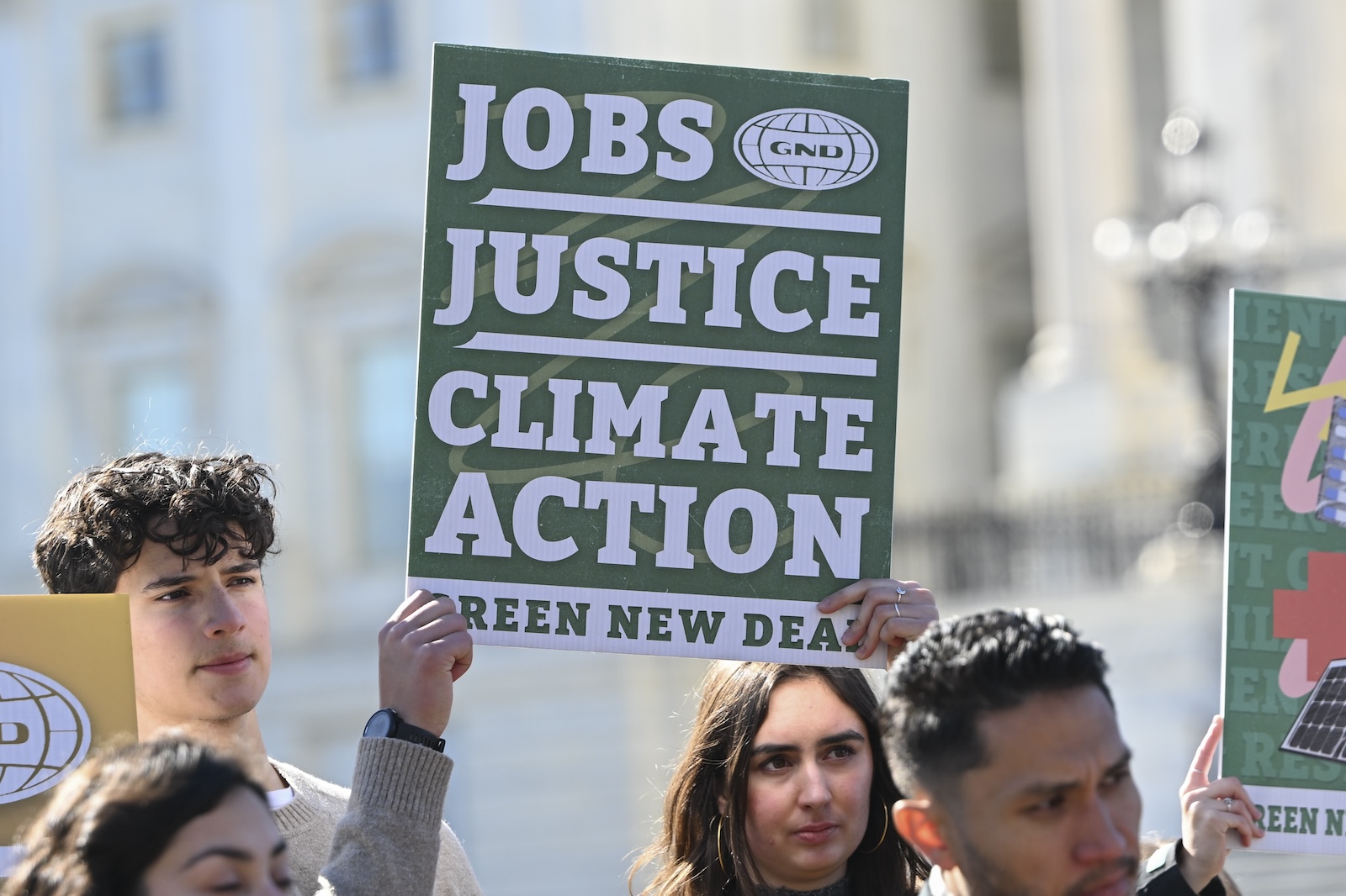 A protest sign saying "Jobs. Justice. Climate Action. Green New Deal" is held up by an audience member at a press conference