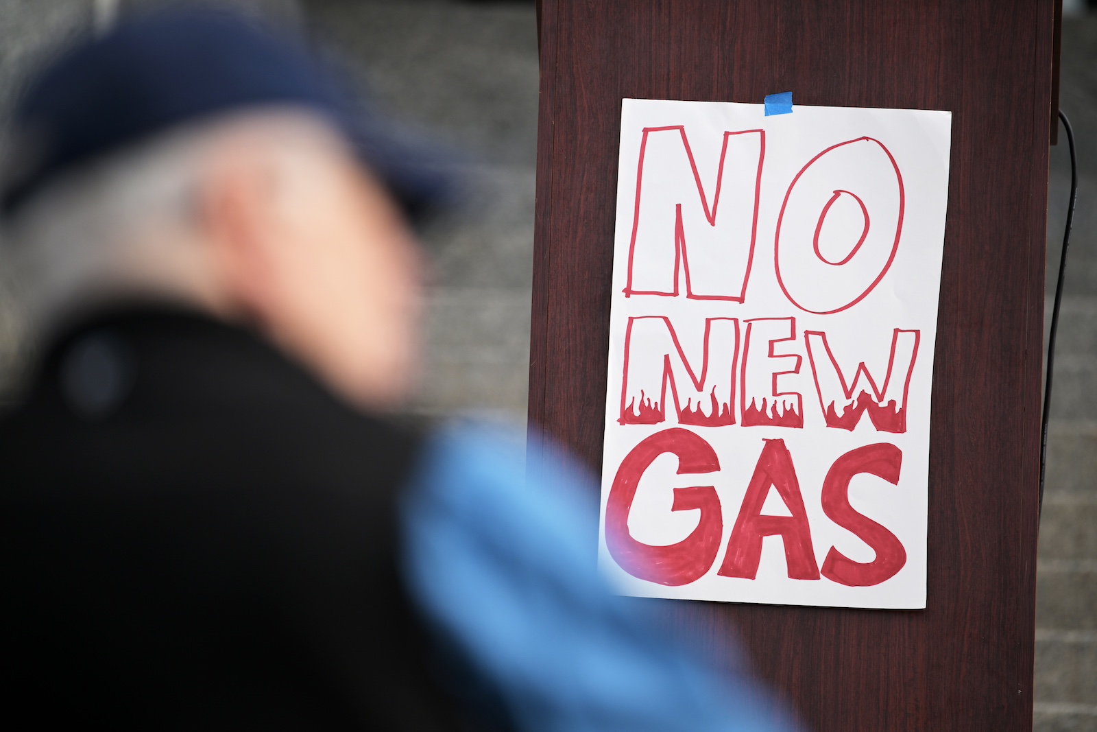 A person's out-of-focus body next to a hand-drawn sign that says 'No New Gas' in red marker on a white poster