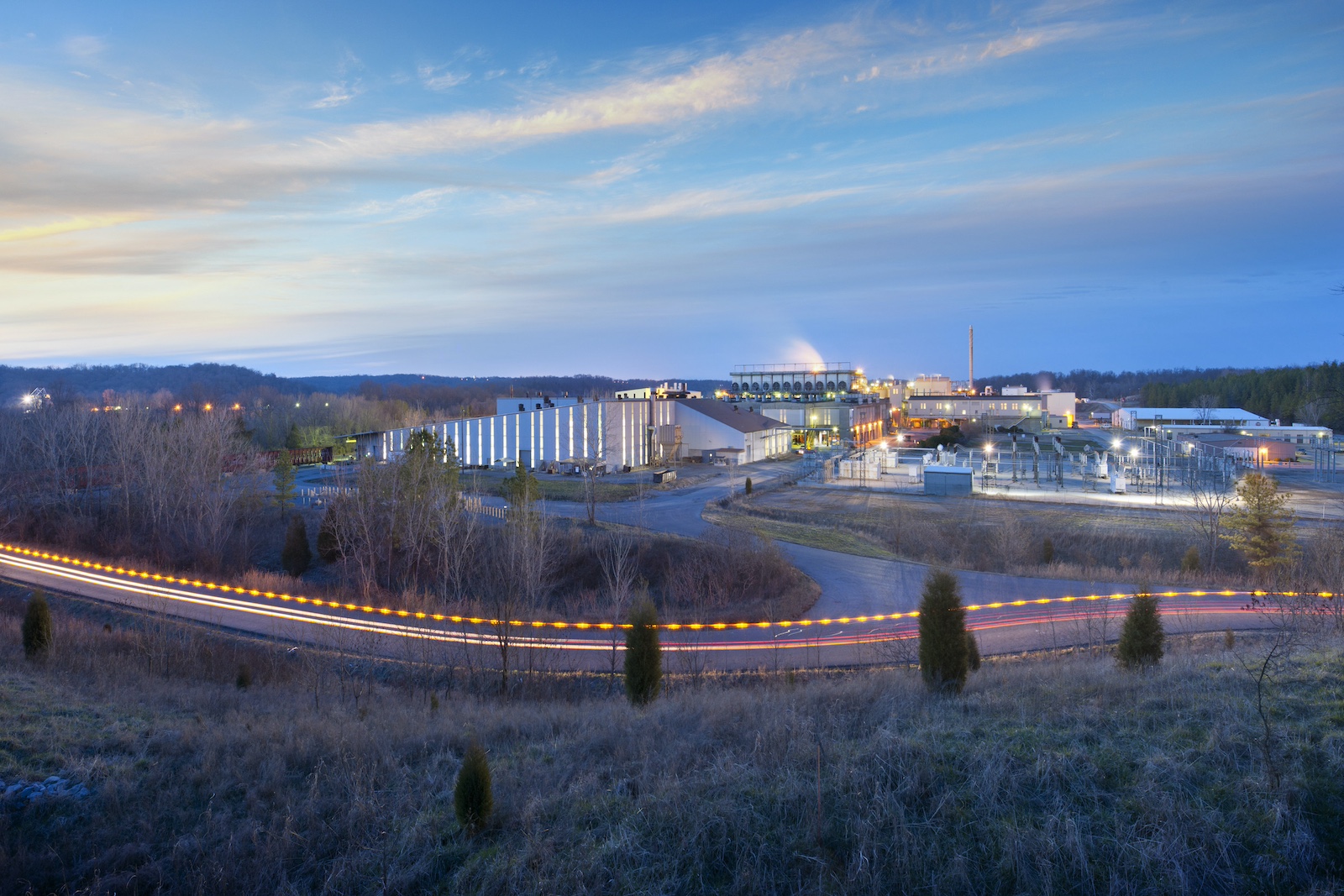 A silvery industrial facility is seen behind some shrubs and a road, with wispy clouds in a blue sky in the background