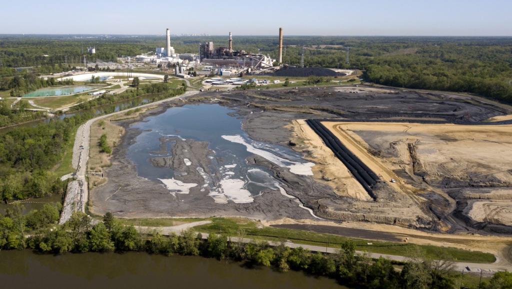 A coal ash pond next to a body of water in foreground. A coal plant in the background and behind it the skyline of the city of Richmond, Virginia.