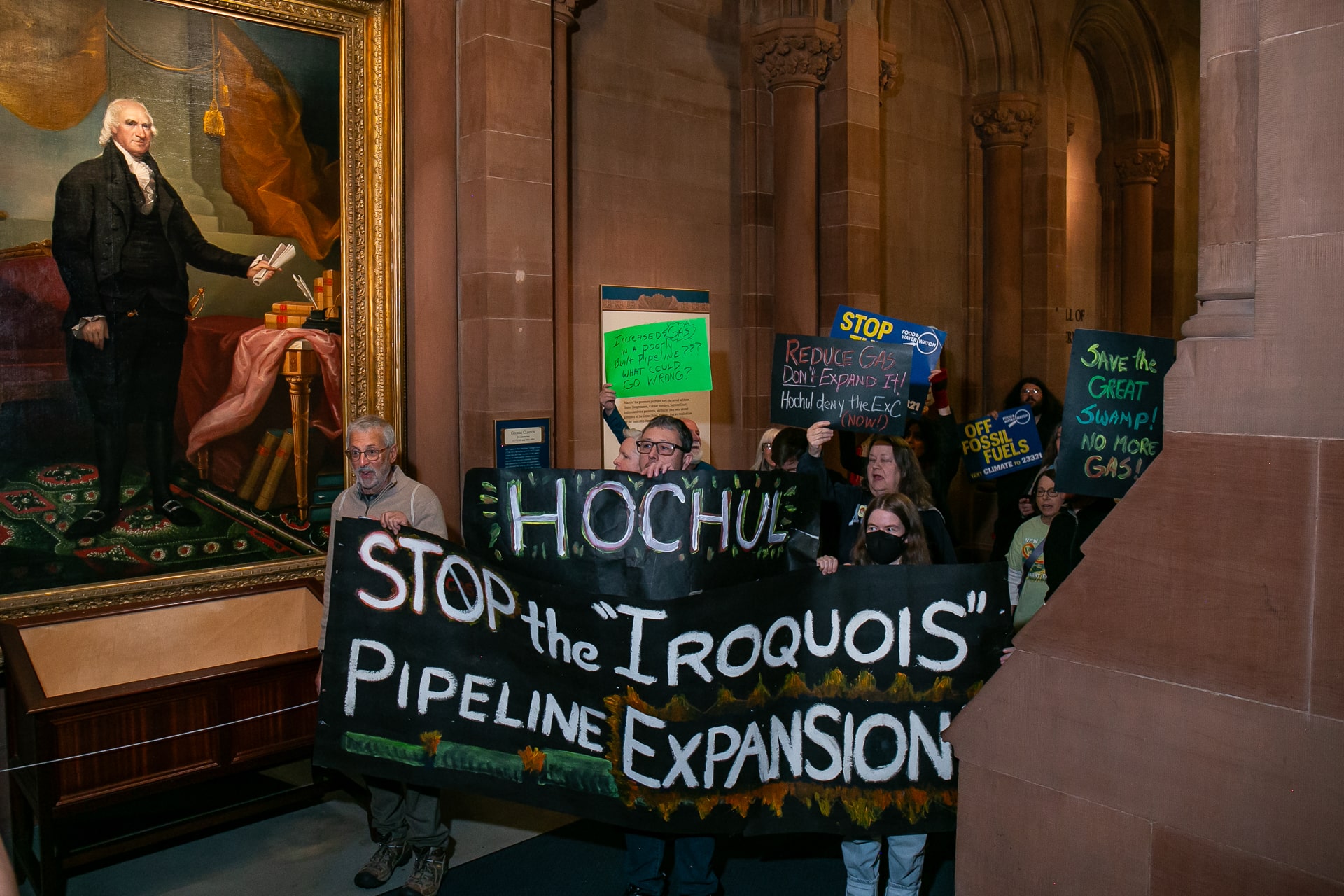 A group of people holding signs that say stop the Iroquois pipeline expansion march in a gilded hallway