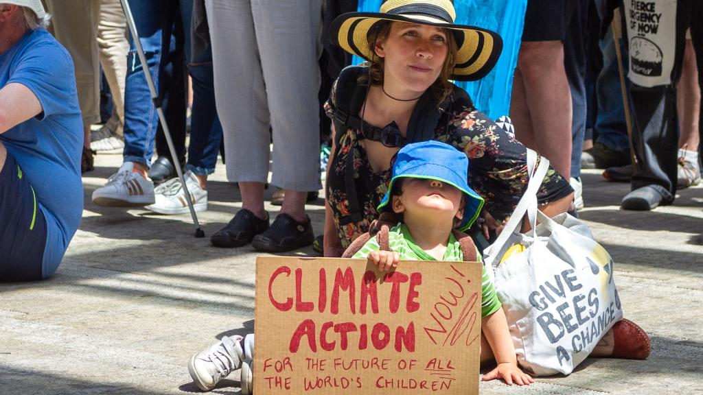A woman sits on the ground with a baby in front of her. They are wearing large hats, and the baby holds a cardboard sign reading 