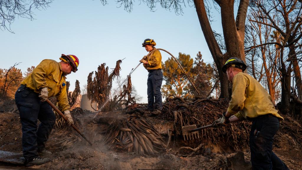 Three firefighters in yellow jackets and red helmets try to put out a fire on a ridge
